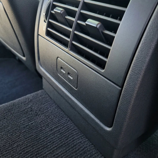 Close-up of a E39 BMW interior showing a black plastic air vent panel with a USB charging port below it, next to a textured gray carpet.