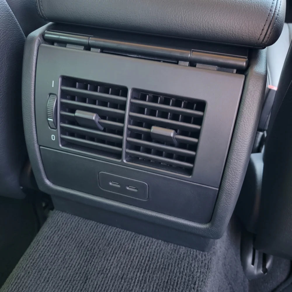 Close-up of a E39 BMW interior showing a black plastic air vent panel with a USB charging port below it, next to a textured gray carpet.